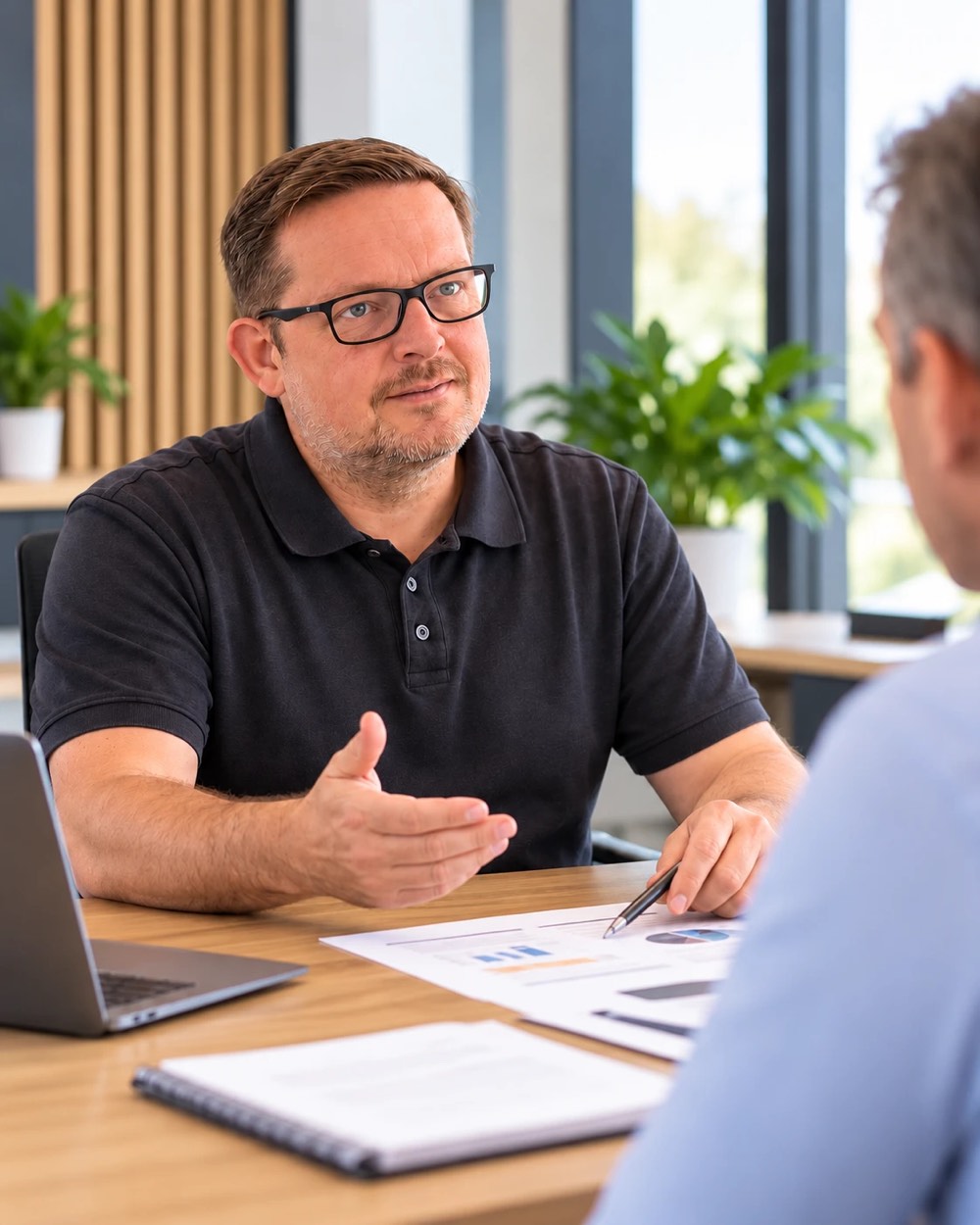 Marc Wilde im schwarzen Poloshirt erklärt einem Kunden am hellen Schreibtisch Unterlagen mit Diagrammen, daneben Laptop und Notizblock
