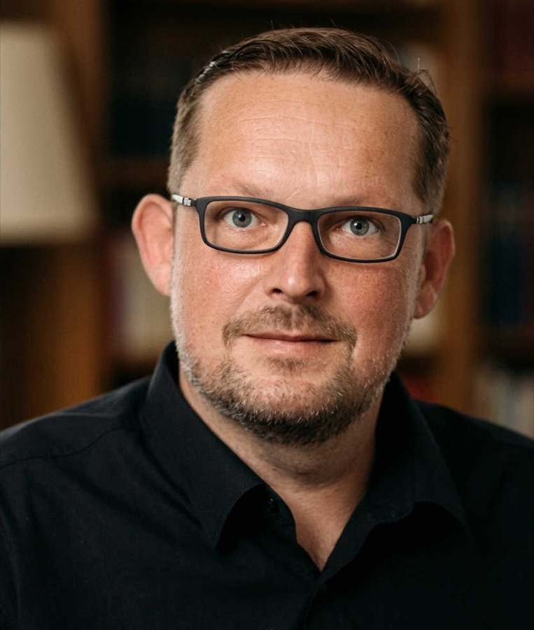 Porträt von Marc Wilde mit dunkler Brille und schwarzem Hemd vor einem unscharfen Bücherregal